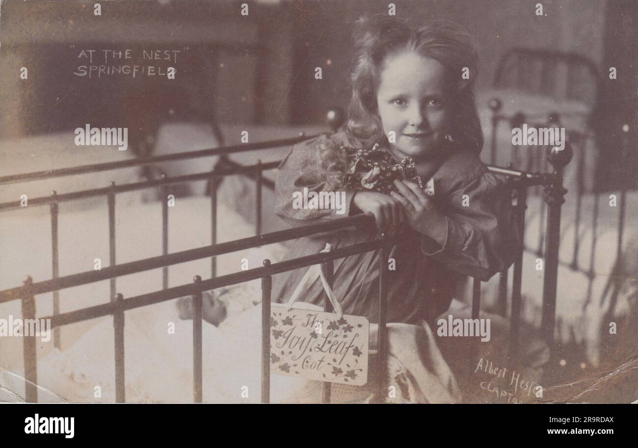 « At the Nest, Springfield » : carte postale photographique du début du 20th siècle. Il montre une jeune fille de l'Armée du Salut diriger la maison d'enfants 'The Nest' au 10 Springfield, Upper Clapton, Londres. Un panneau portant le nom « The Joy Leaf Cot » est accroché aux barres du lit de l'enfant. La carte postale a été envoyée en 1907. Banque D'Images