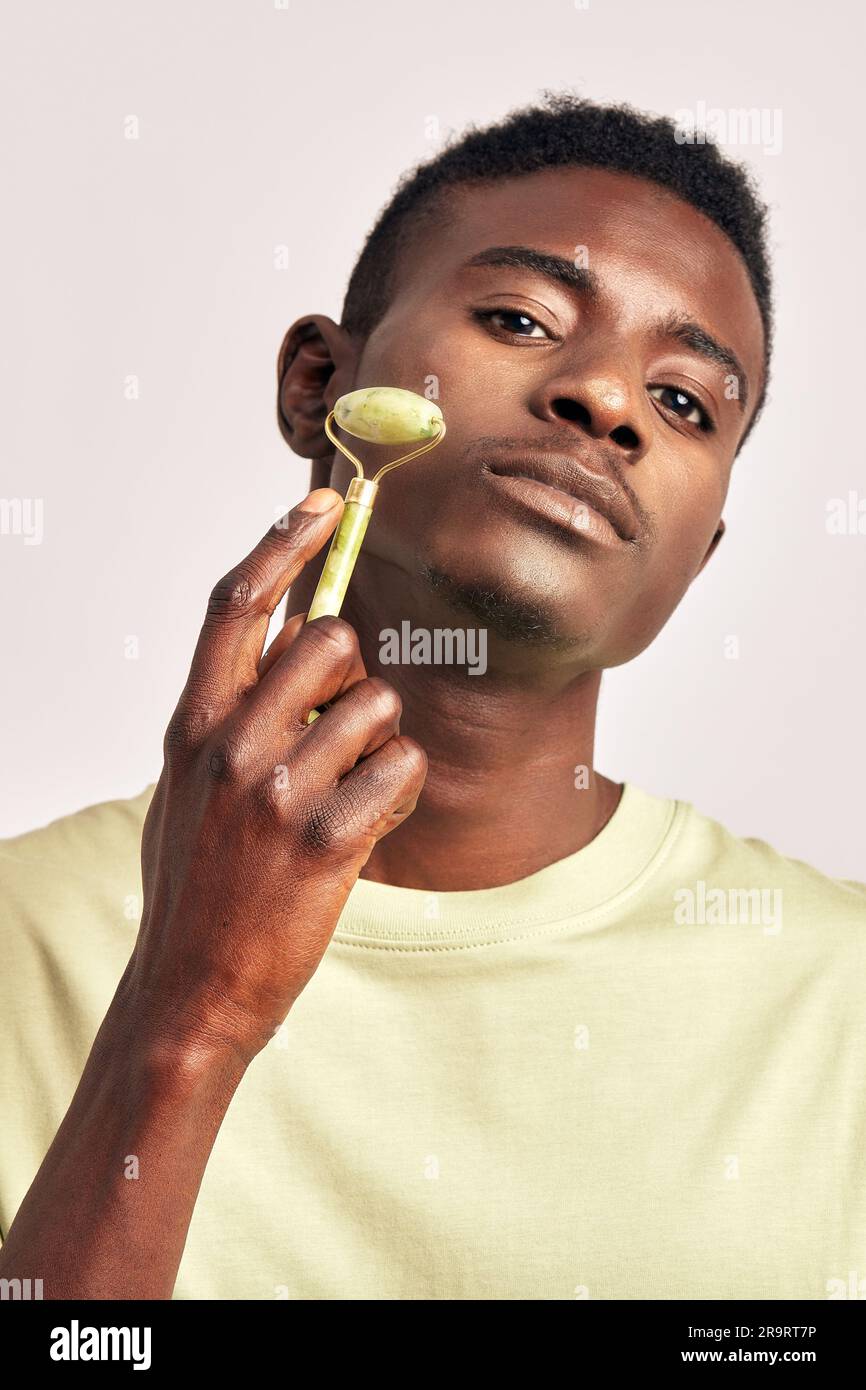 Jeune homme africain, charmant et confiant, utilisant un rouleau de massage du visage jade dans un studio. Un homme noir prenant soin de la peau pour maintenir sa jeunesse et sa santé Banque D'Images