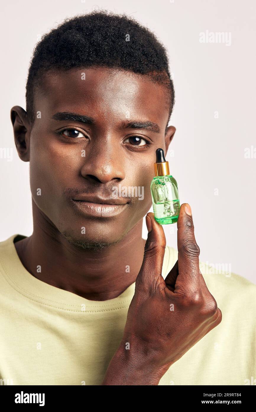 Jeune homme africain charmant et confiant avec un petit flacon de gel hydratant pour le visage dans un studio. Un homme noir prenant soin de la peau pour maintenir son youthfu Banque D'Images