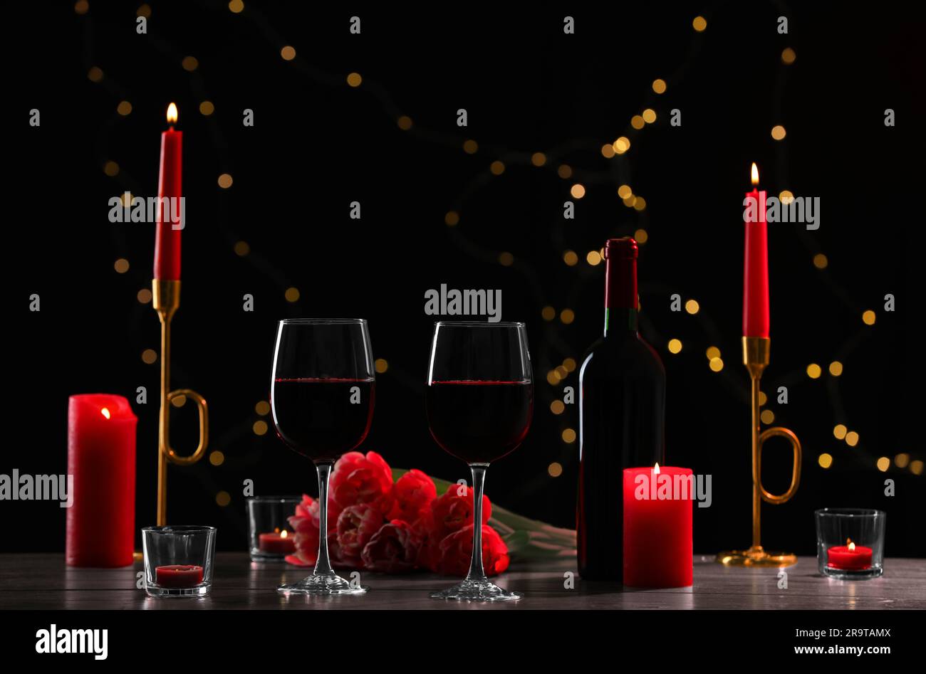 Verres de vin rouge, fleurs de tulipe et bougies allumées sur fond noir contre des lumières floues. Ambiance romantique Banque D'Images