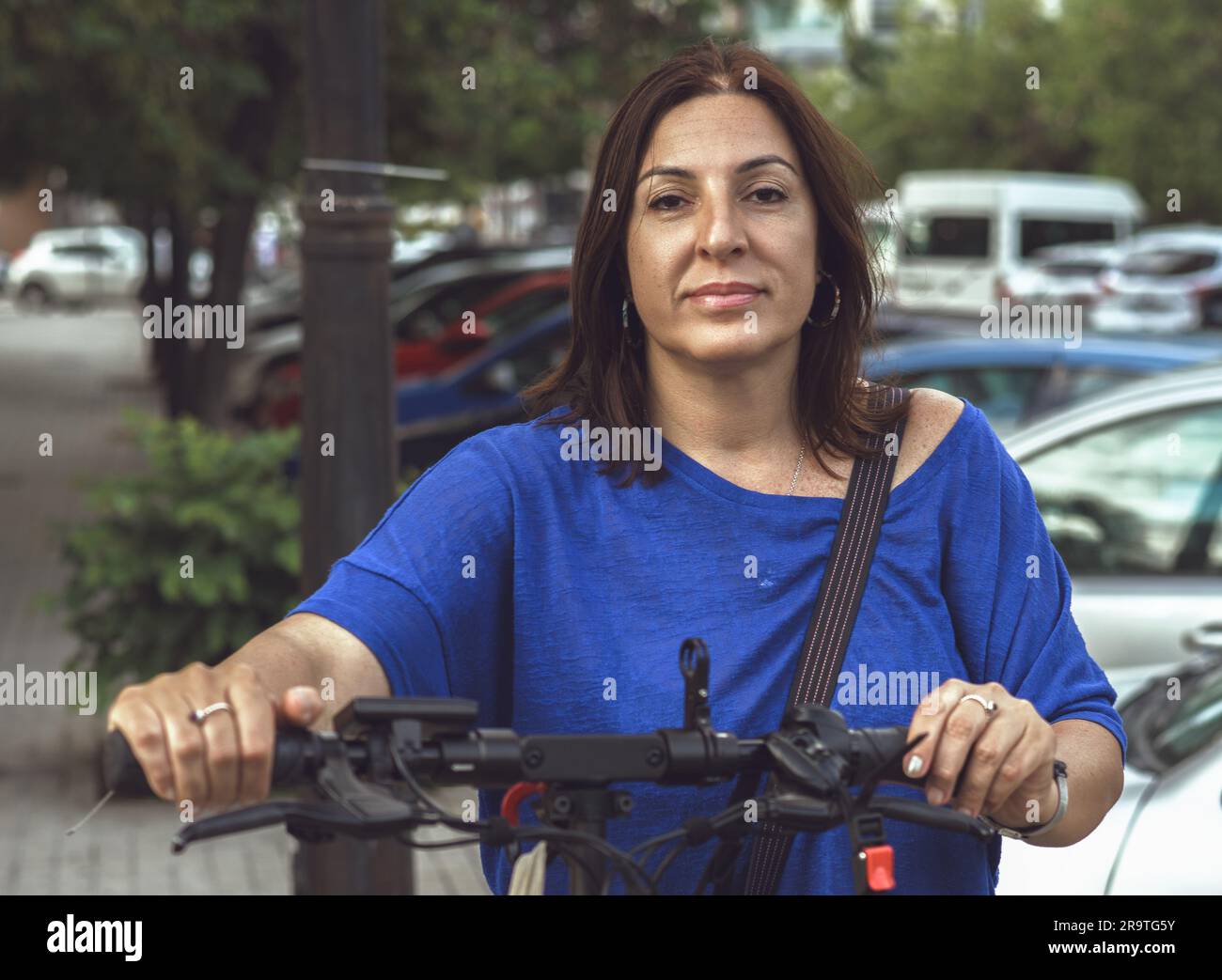 Femme brunette énergique et élégante posant avec son scooter électrique dans un cadre urbain, avec un regard confiant et direct vers la caméra Banque D'Images