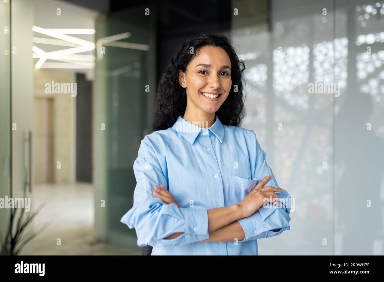 Portrait d'une femme d'affaires heureuse et réussie, patron en chemise souriant et regardant l'appareil photo à l'intérieur du bureau avec des bras croisés, femme hispanique avec boucle Banque D'Images