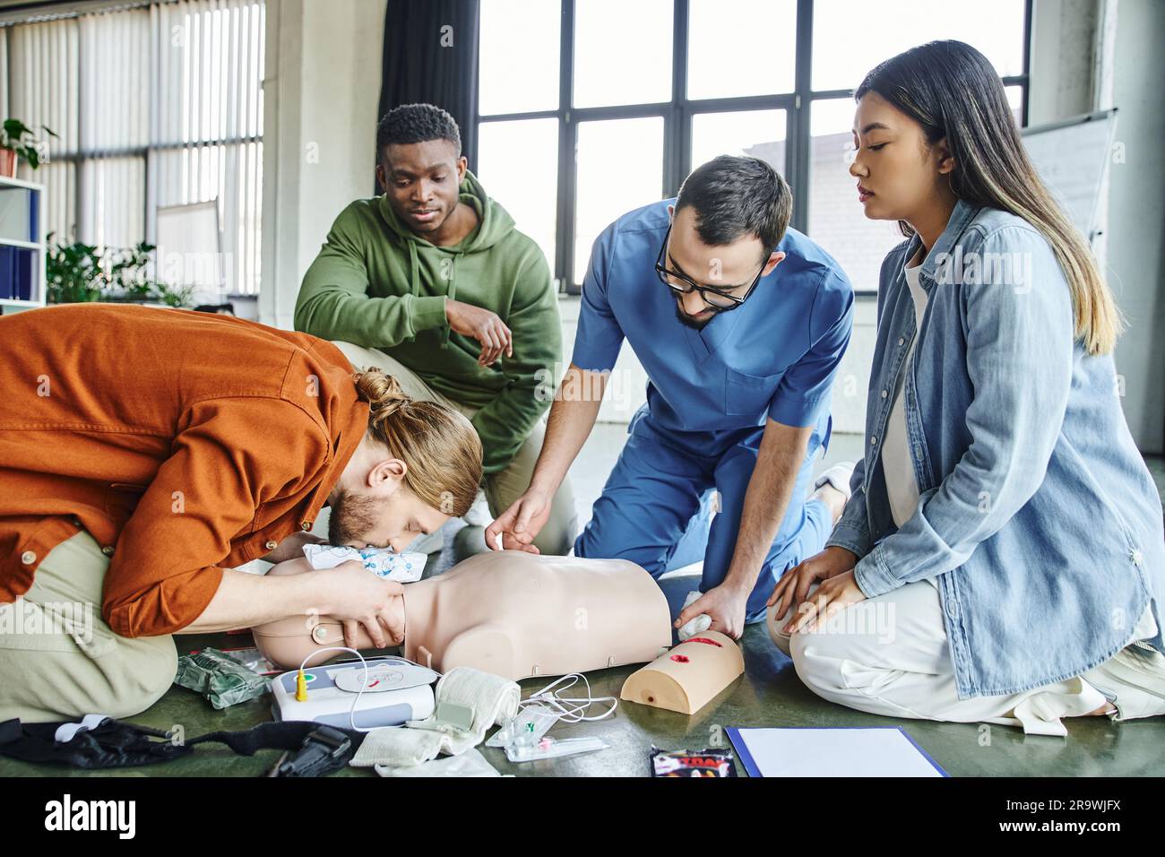 Jeune homme pratiquant la respiration artificielle sur un mannequin de RCP à proximité d'un défibrillateur, d'un équipement médical, d'une équipe multiethnique et d'un membre de l'équipe paramédical pendant l'apprentissage pratique Banque D'Images