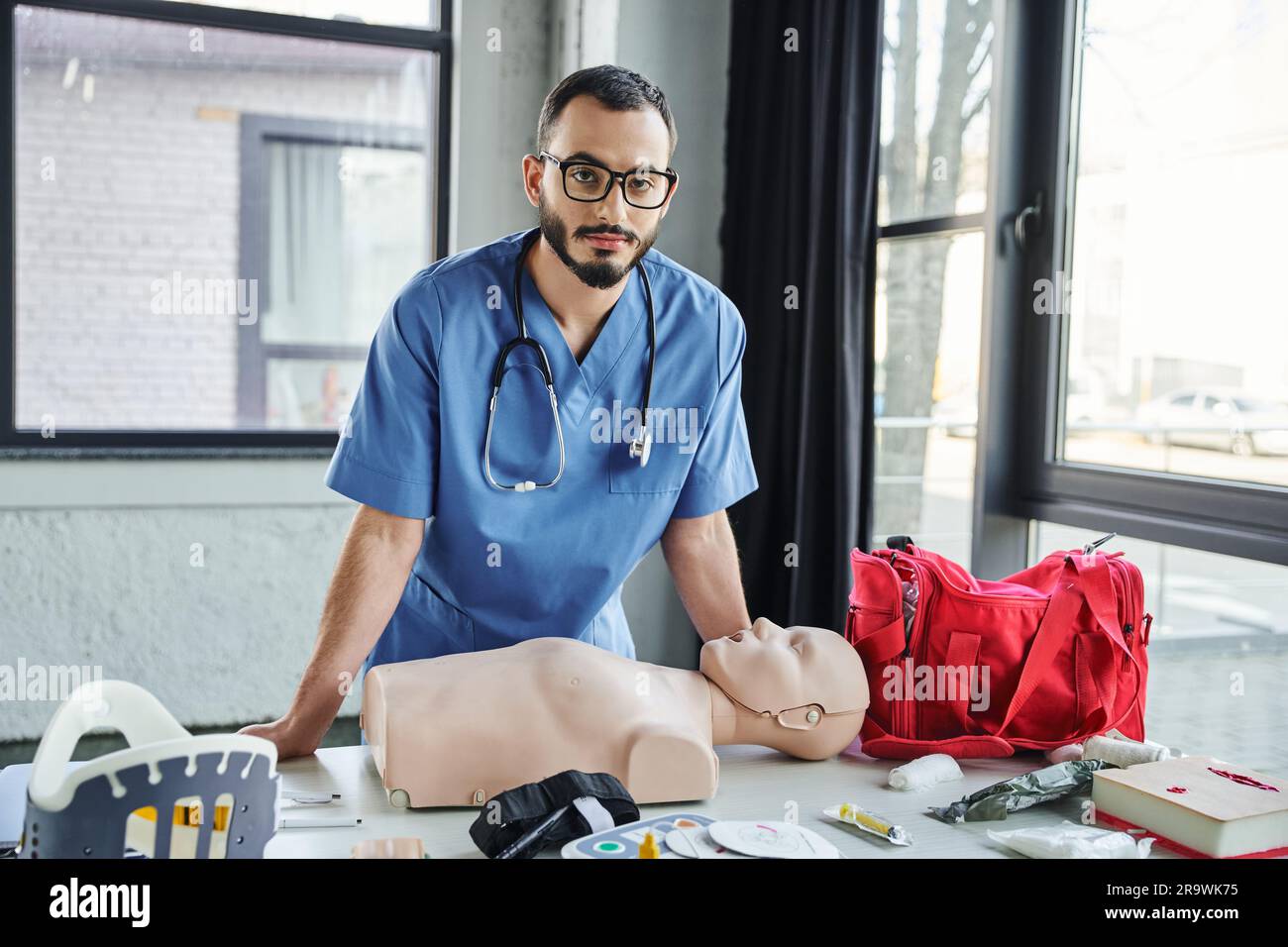 Paramédical barbu dans des lunettes et uniforme bleu regardant la caméra près du mannequin de RCP, sac de premiers secours rouge, défibrillateur et sangle de cou dans la salle de formation, Banque D'Images