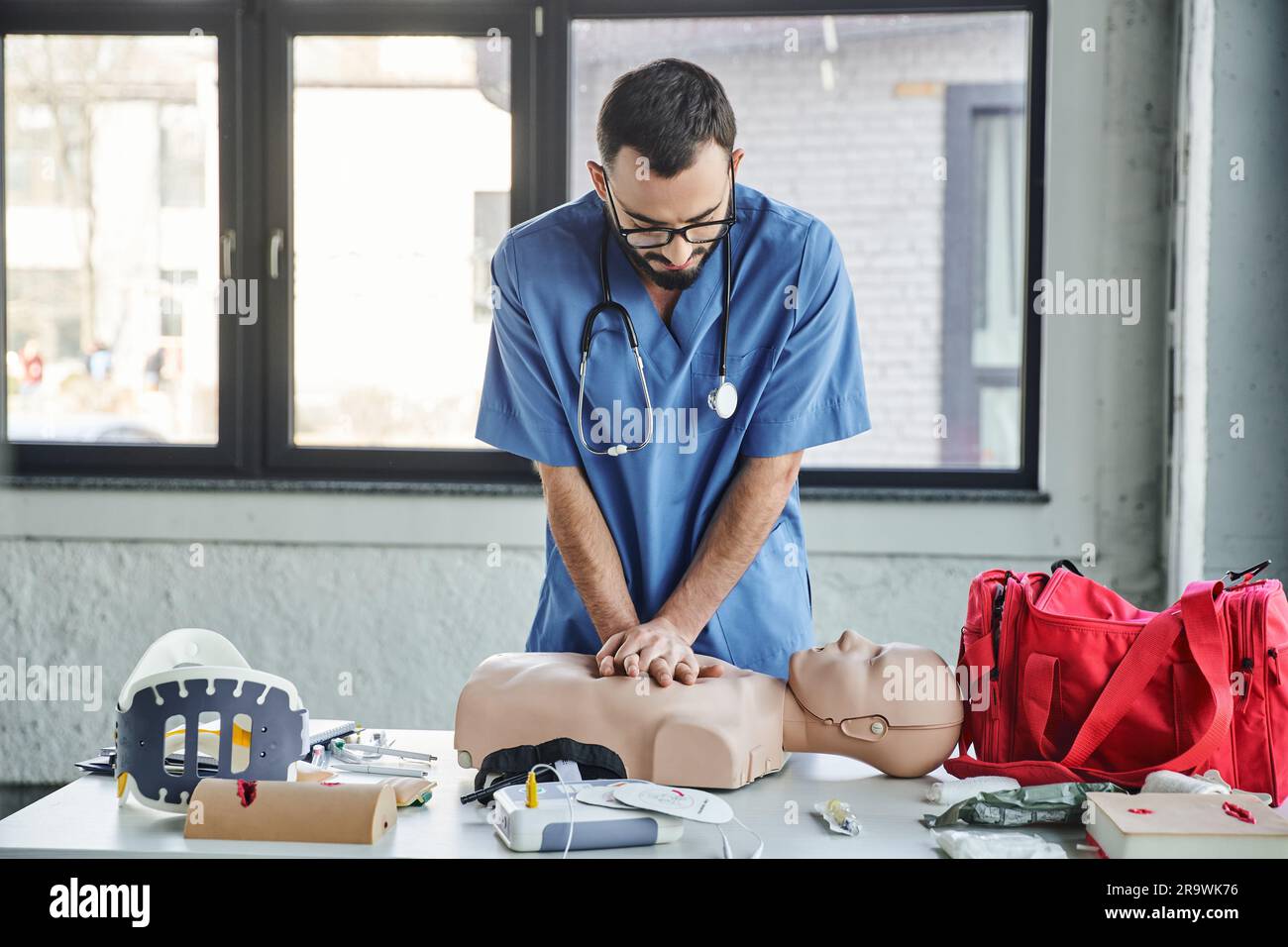 Jeune ambulancier en uniforme bleu et lunettes pratiquant des compressions thoraciques sur un mannequin de RCP près d'un défibrillateur et d'une trousse de premiers soins pendant la semina médicale Banque D'Images