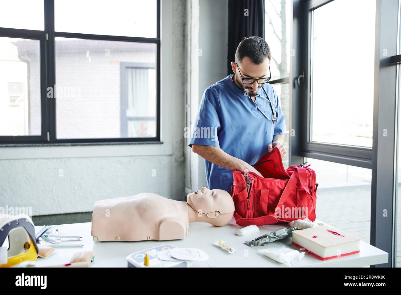 Jeune barbu médecin en uniforme bleu et lunettes de vue déballant le sac de premiers soins rouge près du mannequin de RCP, du défibrillateur automatisé et d'autres équipements en train Banque D'Images