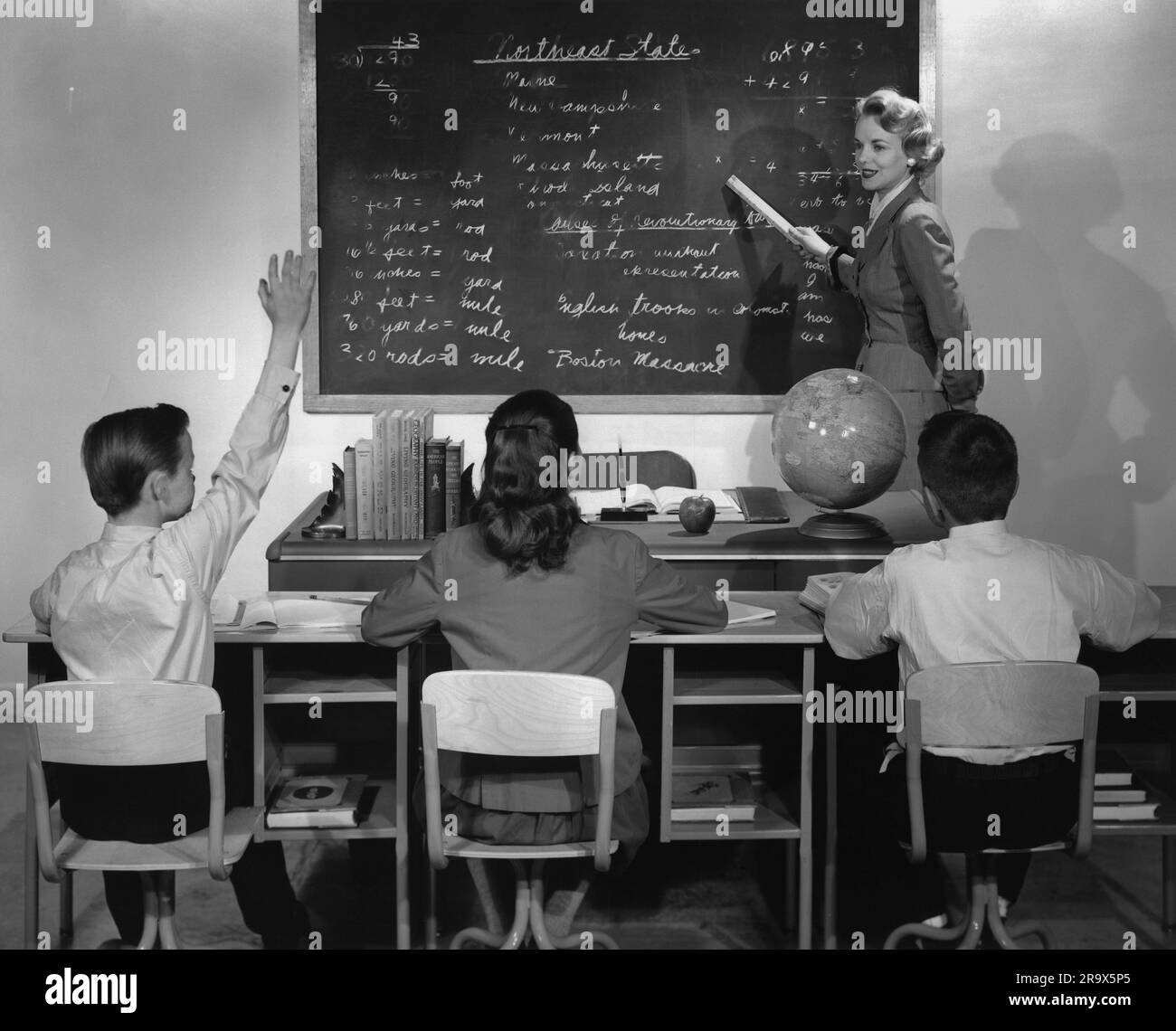 Enseignante debout au tableau noir pointant des écrits à l'aide d'un document enroulé tandis qu'un des trois élèves assis à leur bureau lève la main pour poser une question ou répondre à une question Banque D'Images