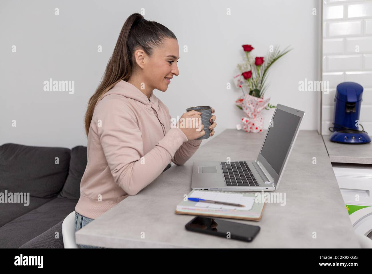 Jeune femme indépendante positive en tenue décontractée avec une tasse de café souriant et parlant par vidéoconférence sur ordinateur portable tout en étant assise à la maison Banque D'Images