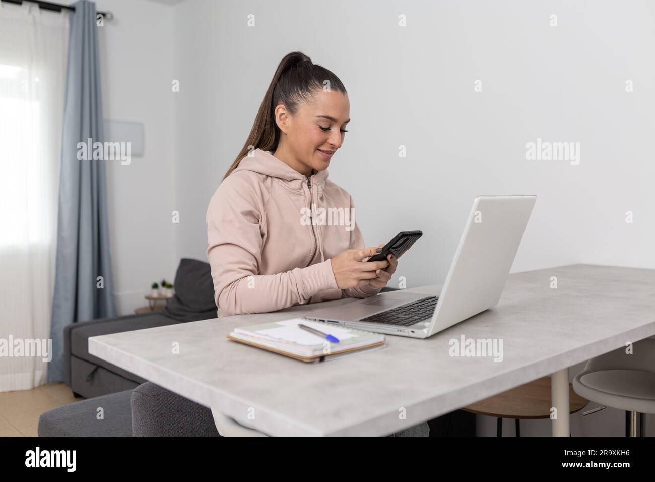 Jeune femme indépendante positive dans des vêtements décontractés parcourant un smartphone tout en travaillant sur un projet à distance sur un ordinateur portable à la table avec un ordinateur portable à la maison Banque D'Images