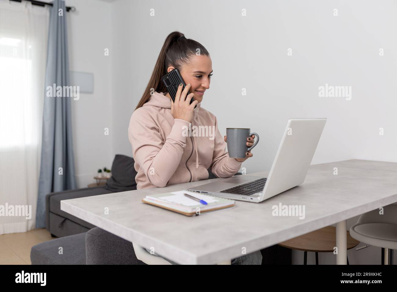 Jeune femme souriante freelance dans des vêtements décontractés ayant conversation téléphonique et boire une tasse de café chaud pendant la pause de travail à distance sur ordinateur portable à Banque D'Images