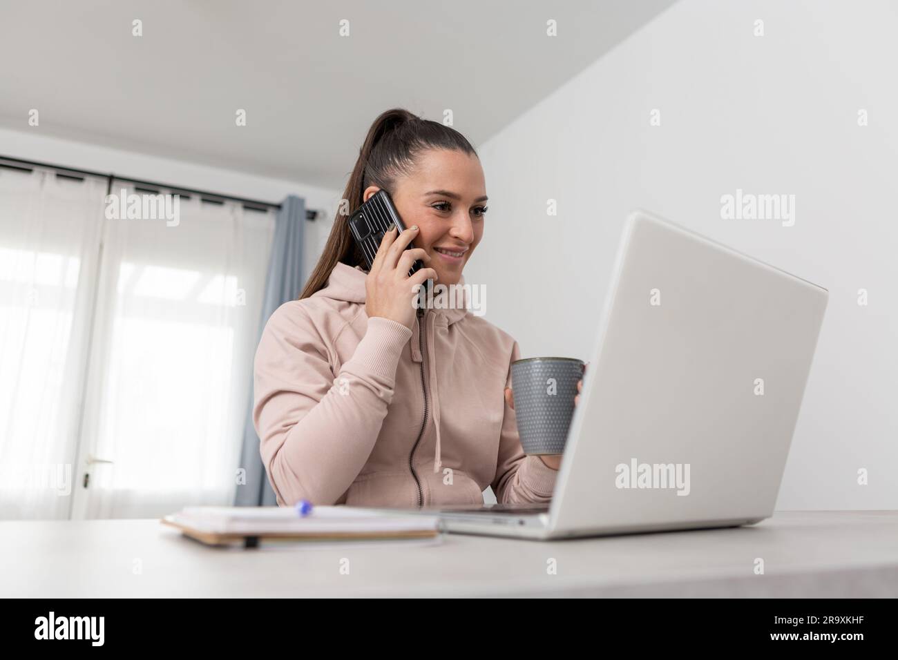 Jeune femme joyeuse freelance à capuche regardant l'écran d'un ordinateur portable et souriant tout en parlant sur un smartphone pendant une pause-café du travail à distance à ho Banque D'Images