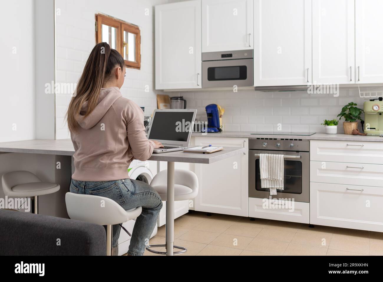 Femme méconnue assise sur son dos dans la cuisine à la maison à l'aide d'un ordinateur portable Banque D'Images