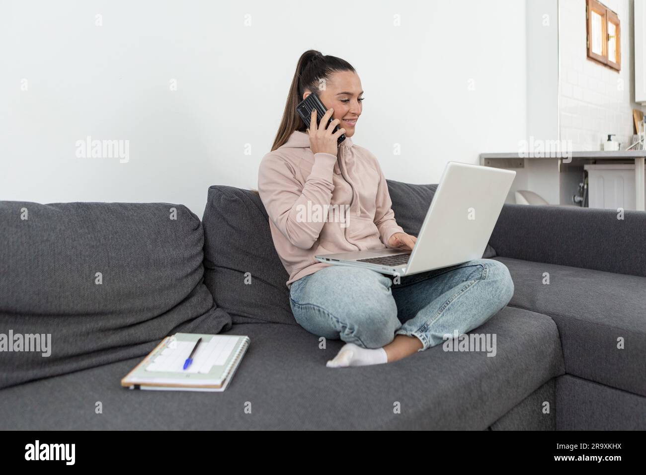 Femme souriante assise sur un canapé en position lotus, parlant au téléphone tout en faisant des achats en ligne avec un ordinateur portable de la maison. Banque D'Images