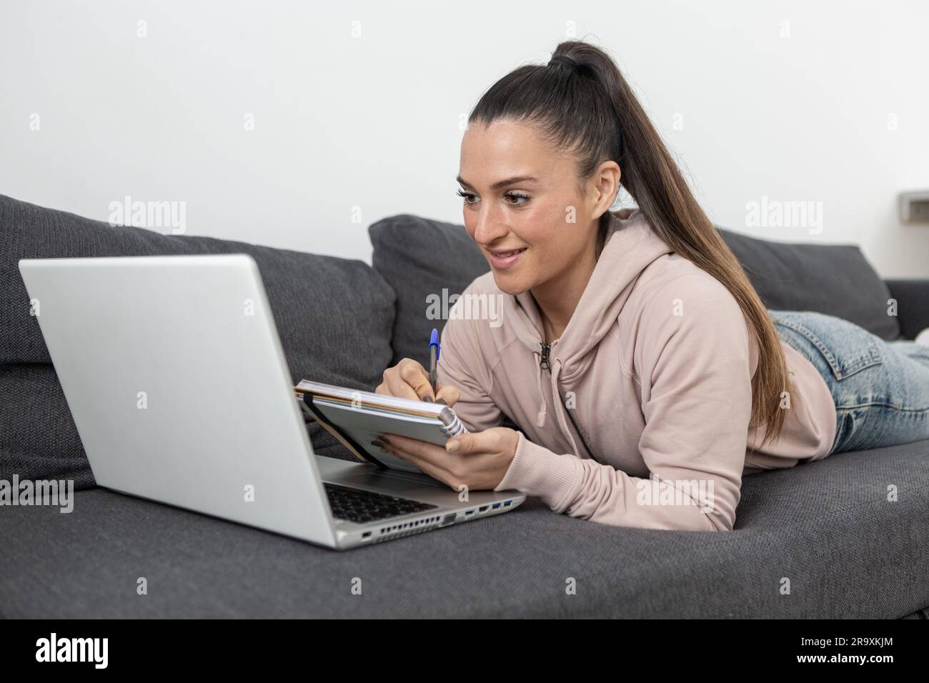 femme vêtue de vêtements décontractés souriant, couchée sur le canapé à la maison, prenant des notes dans un ordinateur portable regardant l'ordinateur portable Banque D'Images
