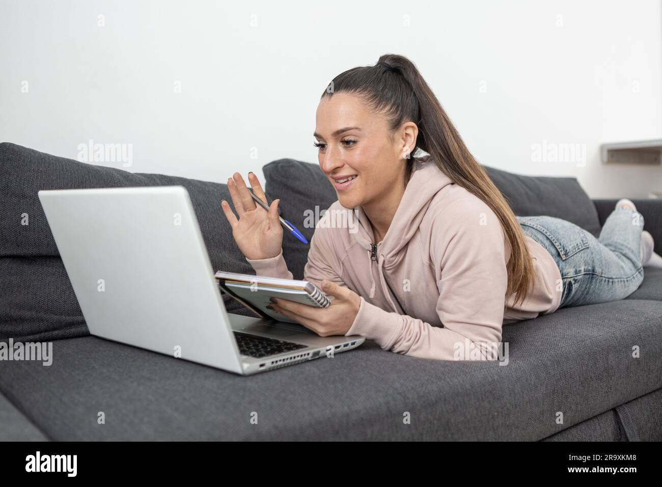 Femme couchée sur le canapé dans des vêtements décontractés souriant, agitant à l'appareil photo de son ordinateur portable tout en prenant des notes dans un ordinateur portable Banque D'Images