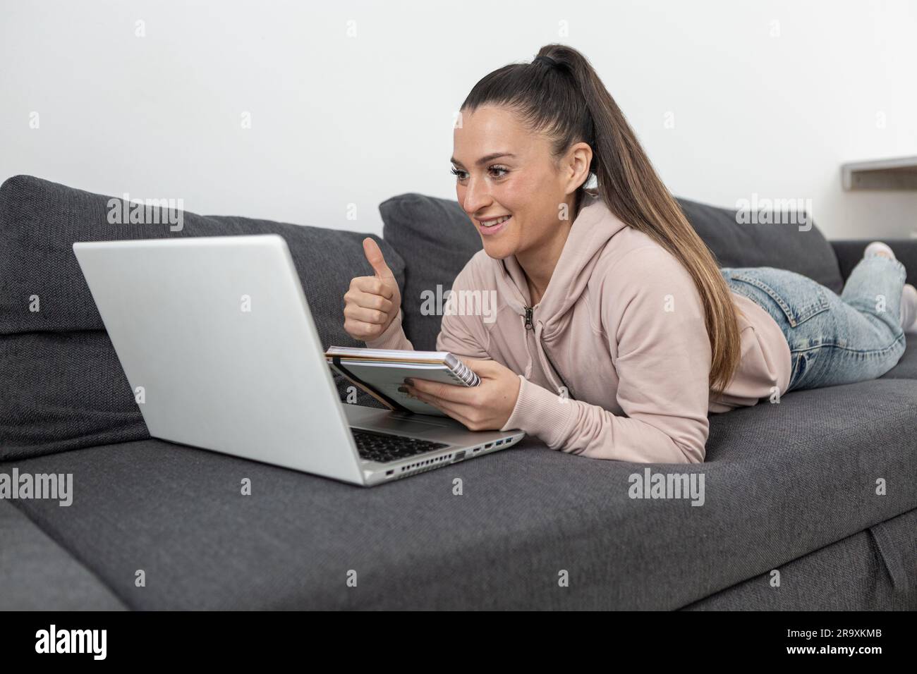 femme souriante dans des vêtements décontractés couchés sur un canapé avec les pouces vers le haut prenant des notes dans un ordinateur portable regardant l'ordinateur portable Banque D'Images