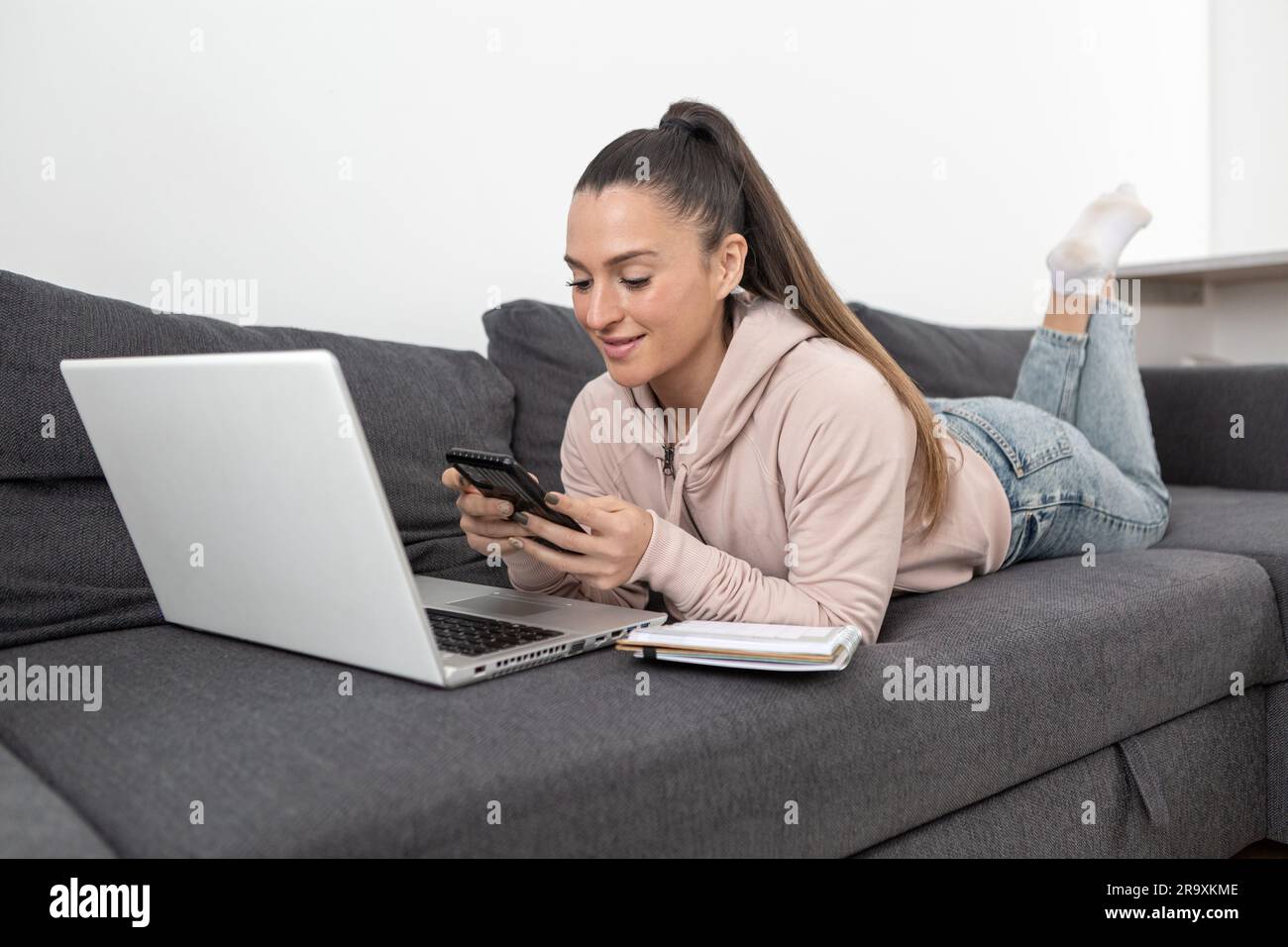 Femme vêtue de vêtements décontractés, couchée sur le canapé à la maison devant l'ordinateur portable tout en regardant son smartphone Banque D'Images