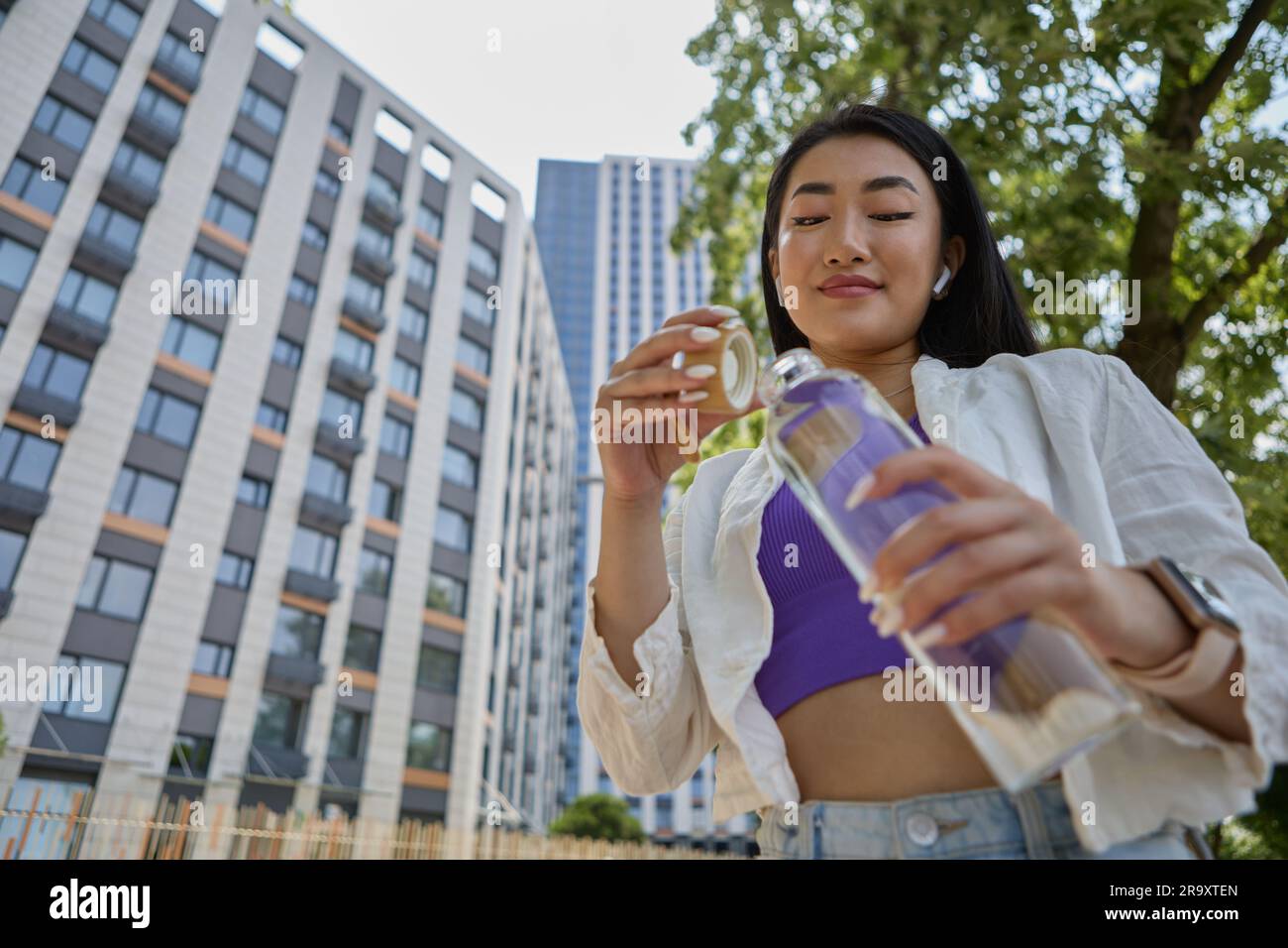 Une femme vietnamienne gaie qui boit de l'eau minérale depuis une bouteille de verre à l'extérieur. Portrait d'une belle jeune femme d'Asie du Sud-est hydratant Banque D'Images