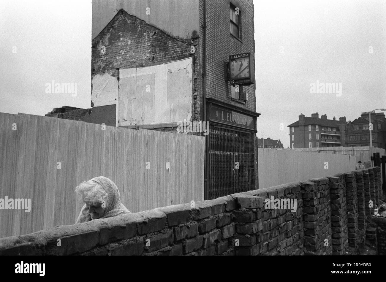 Pauvreté Londres 1970s. Royaume-Uni dans une ruelle au large de Whitechapel High Street, la tête inclinée contre le froid, un résident âgé se bat contre L. Elgrod horloger et bijoutier, qui se tient seul à défier le promoteur immobilier et l'équipe de démolition. Whitechapel, Londres, Angleterre 1975 Royaume-Uni HOMER SYKES Banque D'Images
