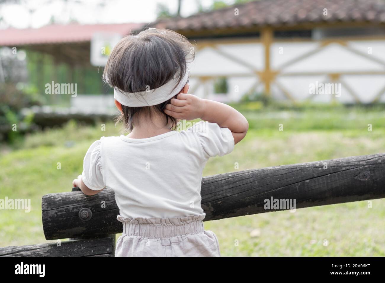 Vue de l'arrière d'une petite fille brune debout à côté de la clôture en bois, touchant sa tête tout en regardant la nature Banque D'Images
