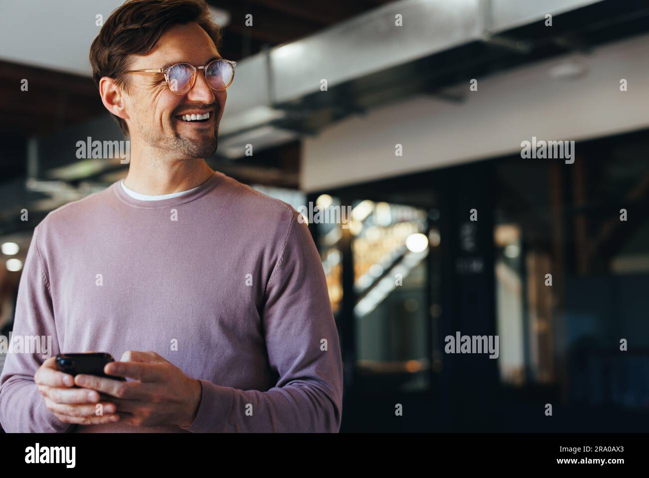 Homme d'affaires pensif souriant à l'aide d'un téléphone portable dans un lieu de travail moderne. Un homme d'affaires heureux qui envoie des SMS sur un téléphone portable lorsqu'il se trouve dans un bureau. Banque D'Images