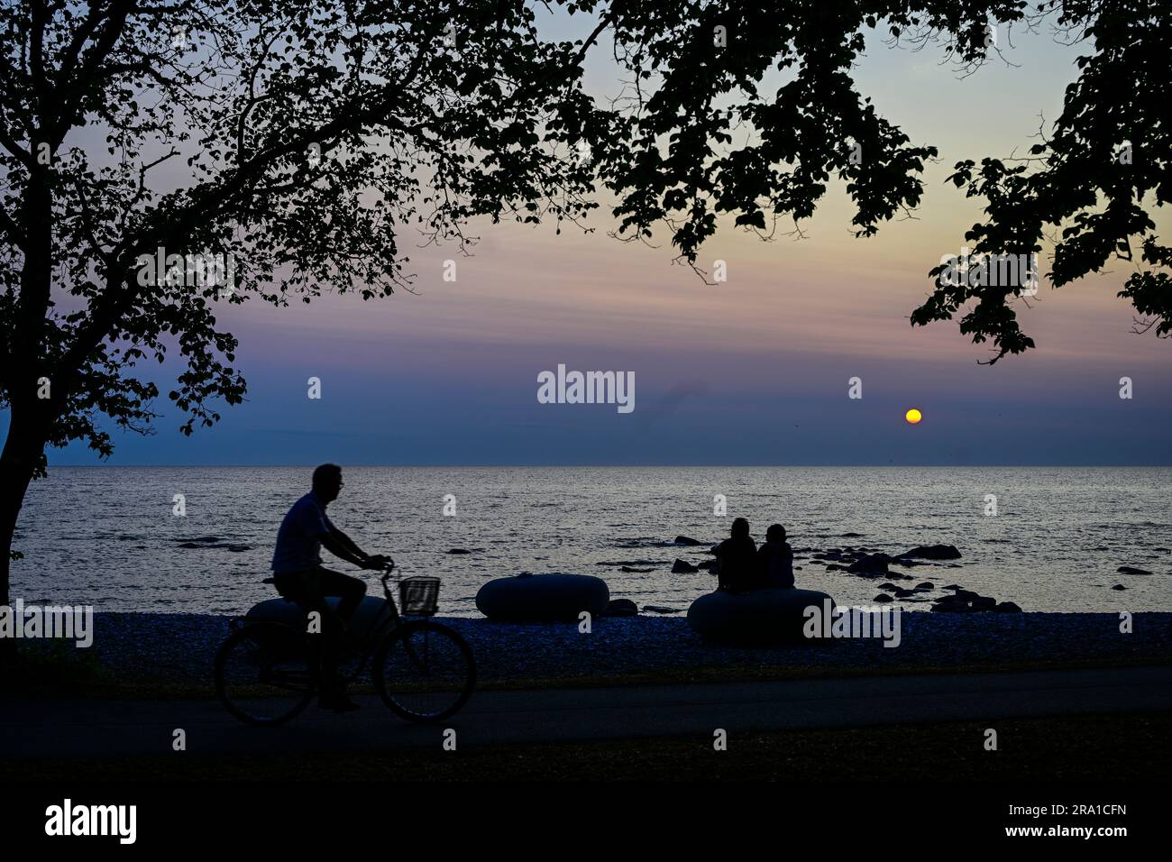 Une personne à vélo par deux personnes assises sur une grande pierre sur la plage en profitant du coucher du soleil à Visby, Suède, 29 juin 2023. Photo: Anders Wiklund / TT / Banque D'Images