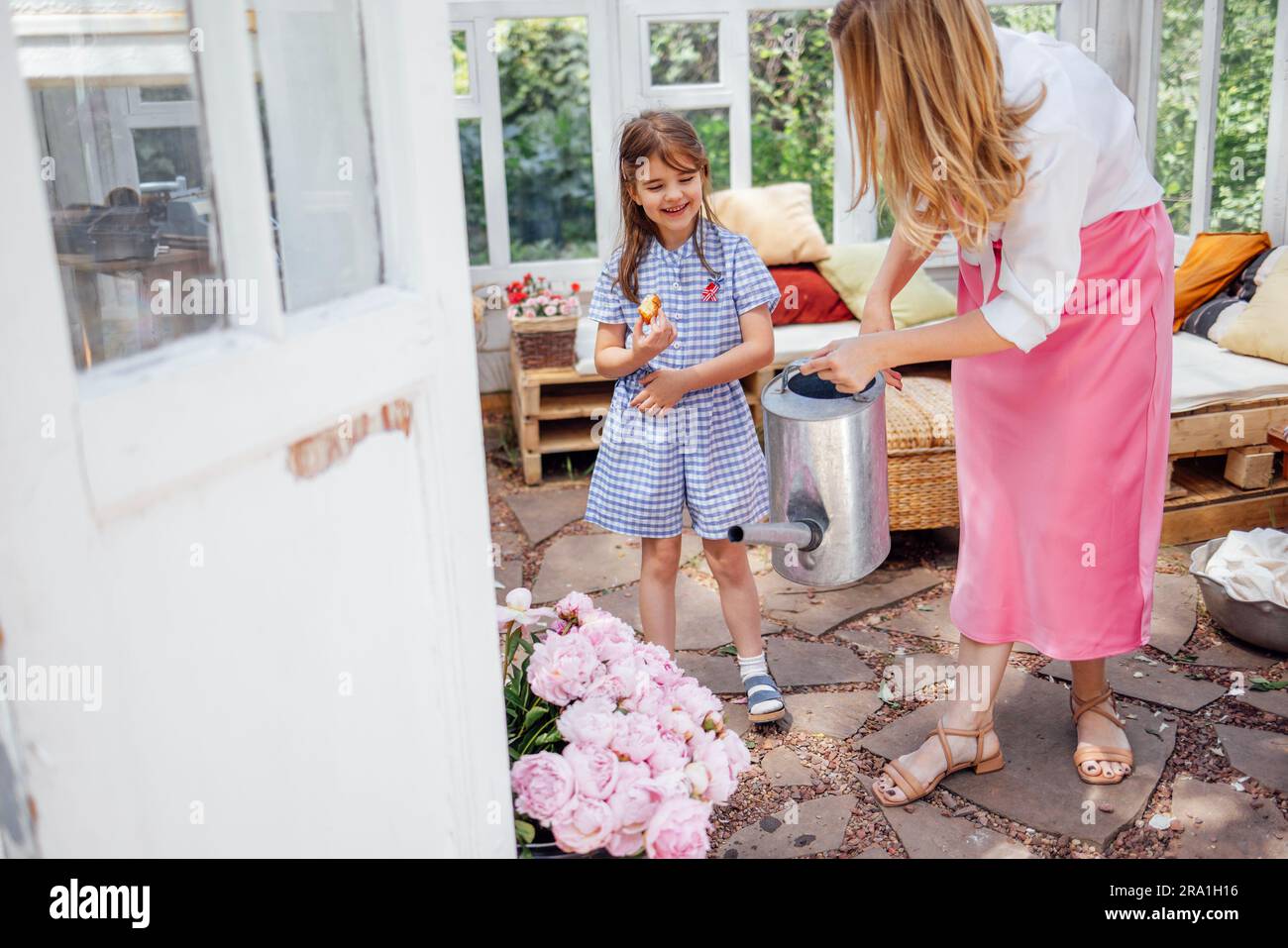 Maman blonde avec sa petite fille riante est arroser des pivoines d'un arrosoir dans une maison d'été. Une jeune femme en robe rose et sa jolie fille Banque D'Images