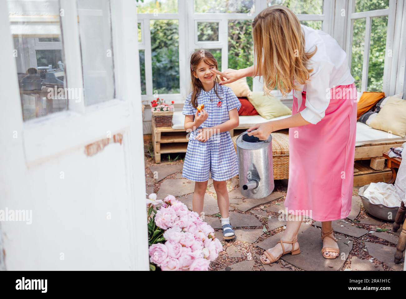 Maman blonde avec sa petite fille riante est arroser des pivoines d'un arrosoir dans une maison d'été. Une jeune femme en robe rose et sa jolie fille Banque D'Images