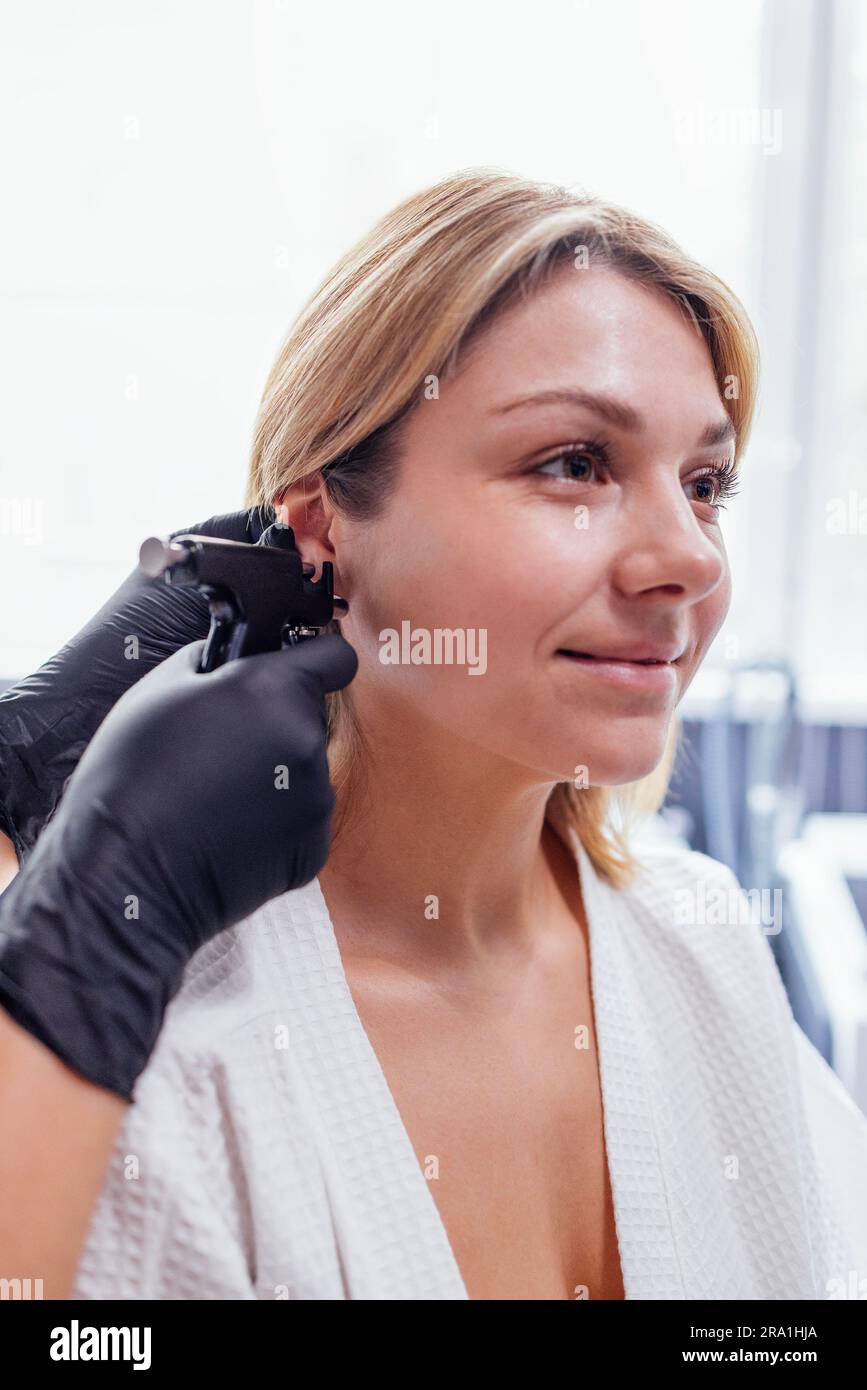 Gros plan sur le visage d'une jeune femme blonde effectuant une procédure de perçage d'oreilles. Les mains du médecin en gants noirs tiennent le pistolet de perçage d'oreilles. Travailleur médical faisant trou an Banque D'Images