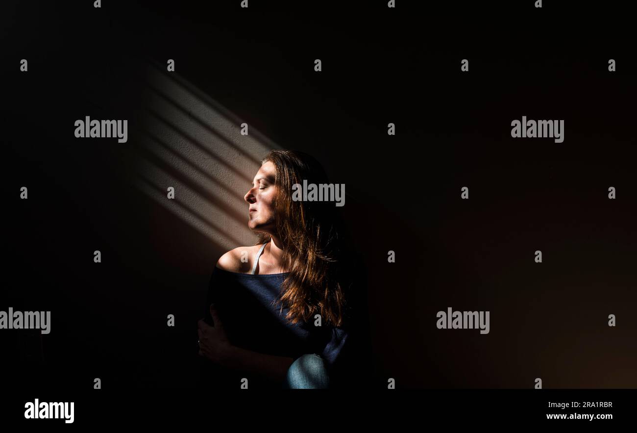 Profil d'une femme assise dans une zone de lumière dans une pièce sombre. Banque D'Images