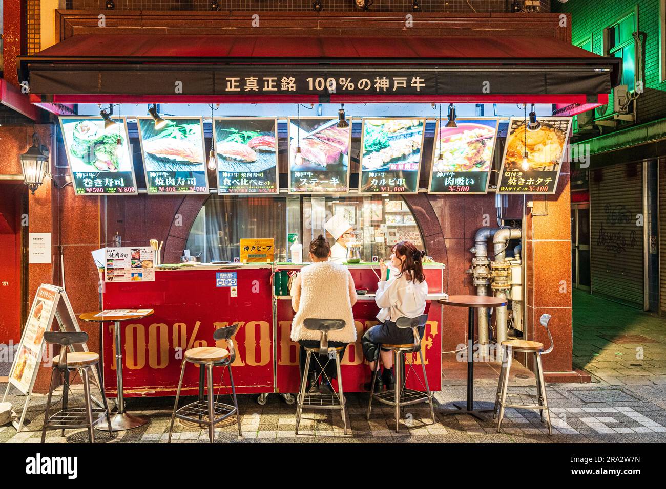 Deux jeunes femmes mangeant avec des baguettes tout en étant assis sur des tabourets dans un bar de cuisine de rue chinois servant du bœuf Kobe, nuit, Chinatown à Kobe, Japon. Banque D'Images