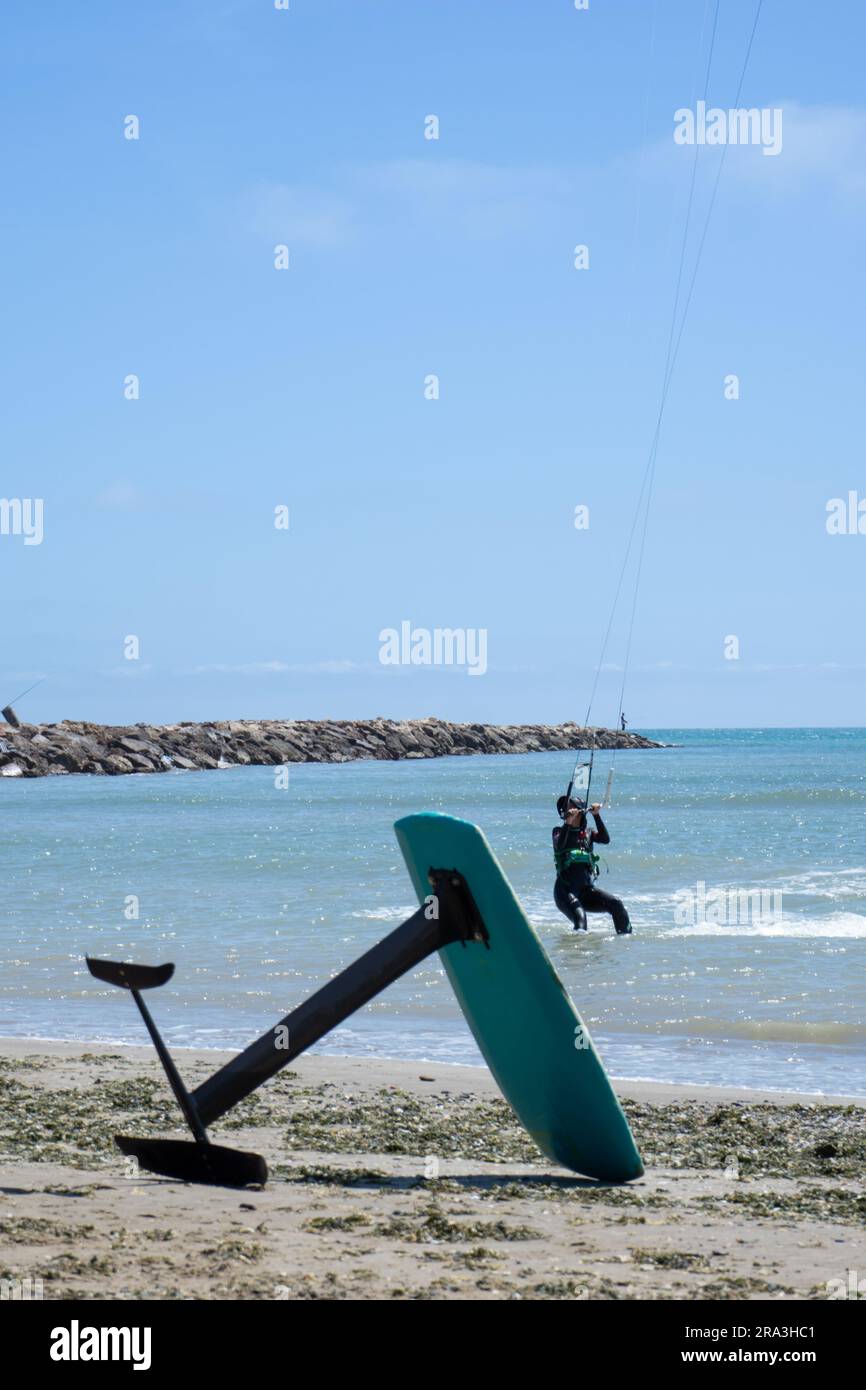 Une planche à feuilles de kitesurf reposant sur le sable de la plage avec une femelle kitesurf dans l'eau en arrière-plan. Concept de sport nautique Banque D'Images