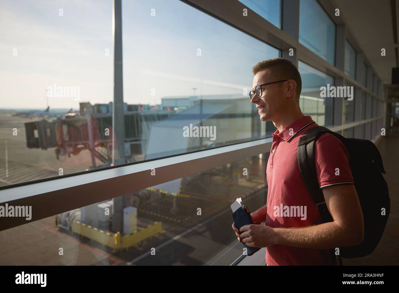 Homme souriant voyage en avion. Passager regardant par la fenêtre sur la piste de l'aéroport tout en tenant son portefeuille avec la carte d'embarquement dans sa main. Banque D'Images