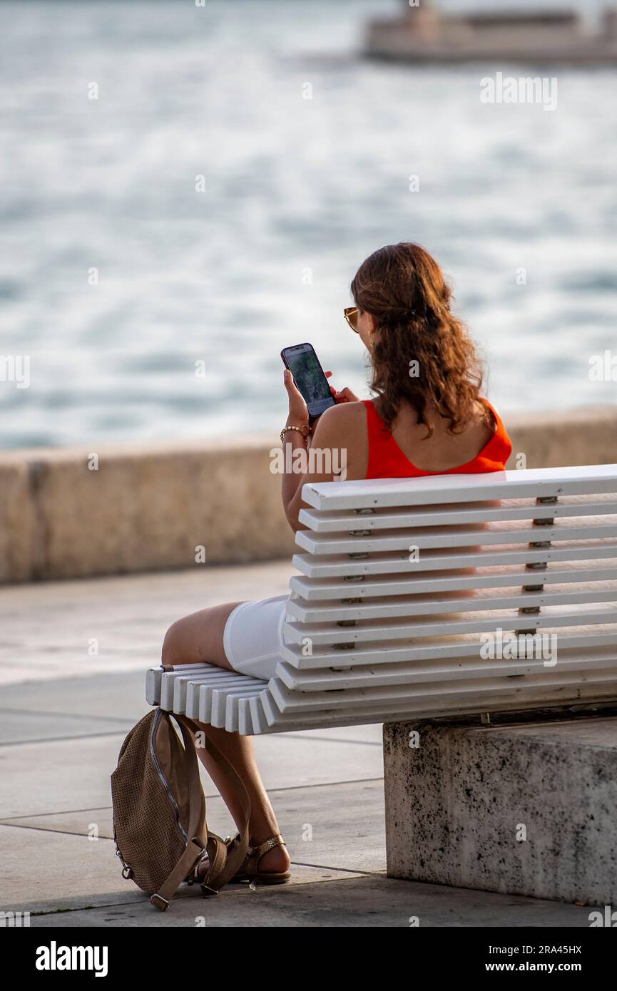 Jeune femme assise au port de bord de mer dans grad Split sur un banc peint en blanc à l'aide d'un smartphone dans le chaud soleil de soirée d'été méditerranéen Banque D'Images