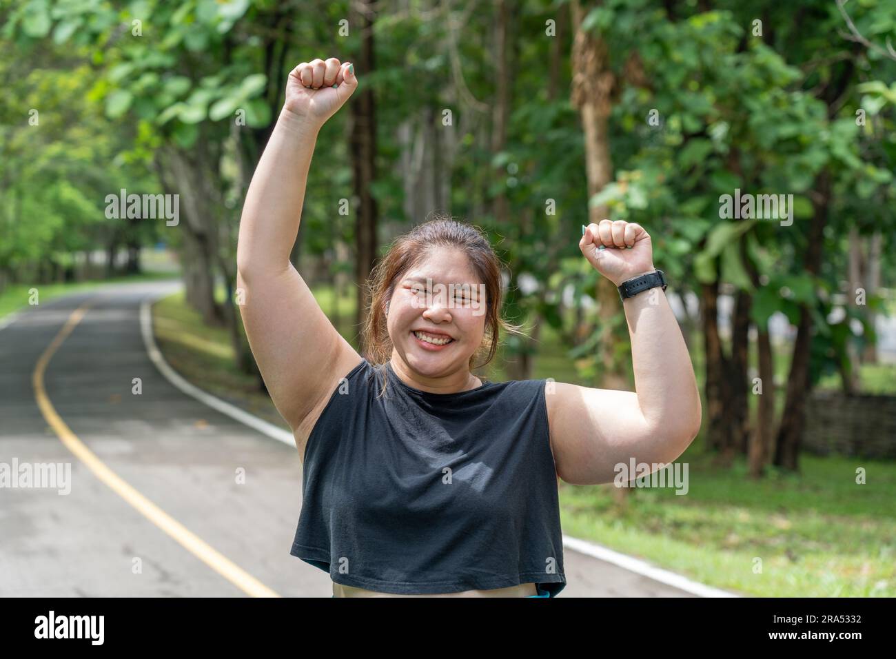 Bonne jeune femme en surpoids souriant et levant les deux mains dans les airs après avoir terminé avec succès son entraînement matinal sur une piste de course d'un lo Banque D'Images