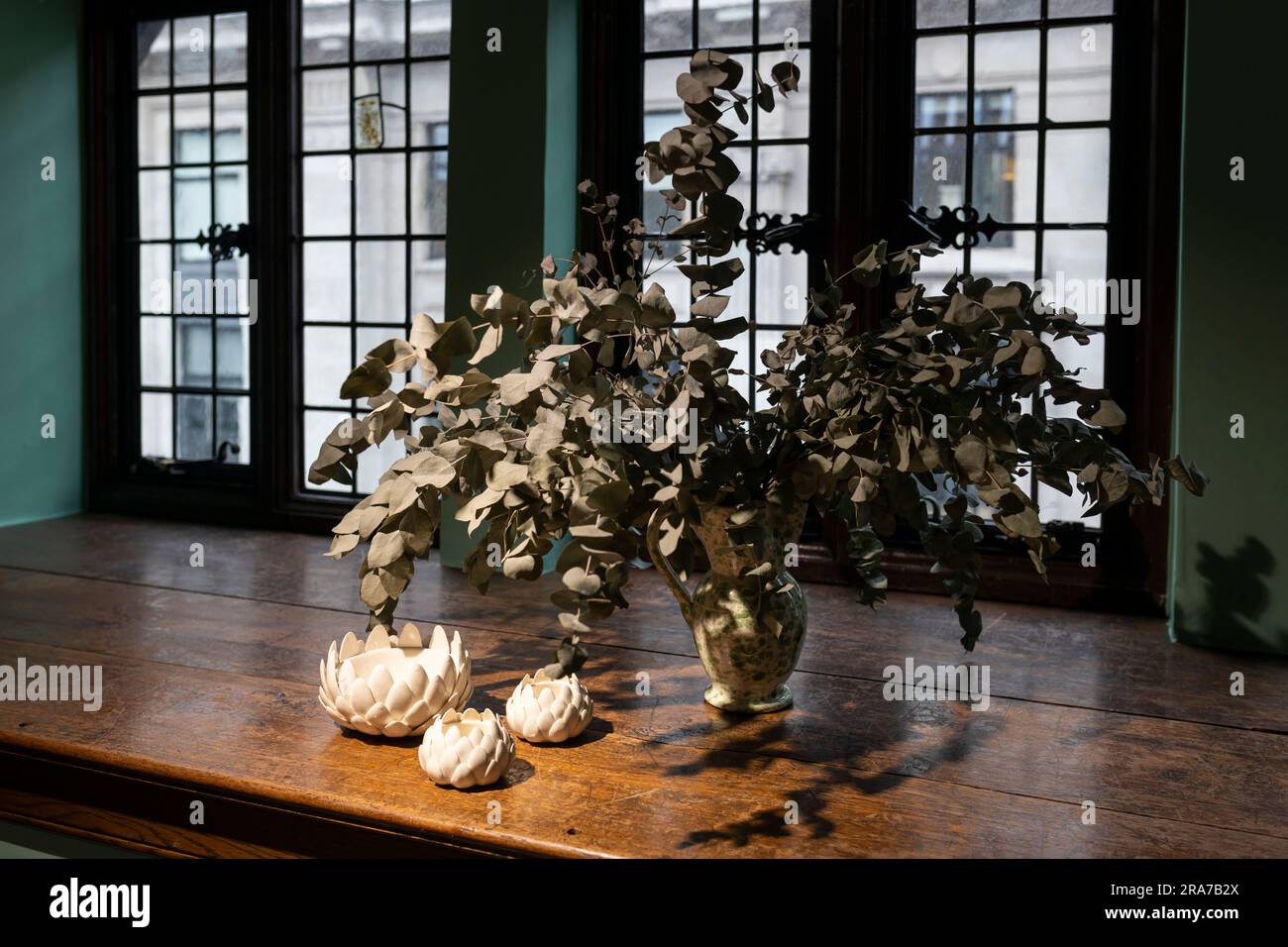 Bouquet de branches artificielles d'eucalyptus dans un pot en céramique à pois avec des figurines d'artichaut en porcelaine blanche sur un large rebord de bois de chêne contre la ba Banque D'Images