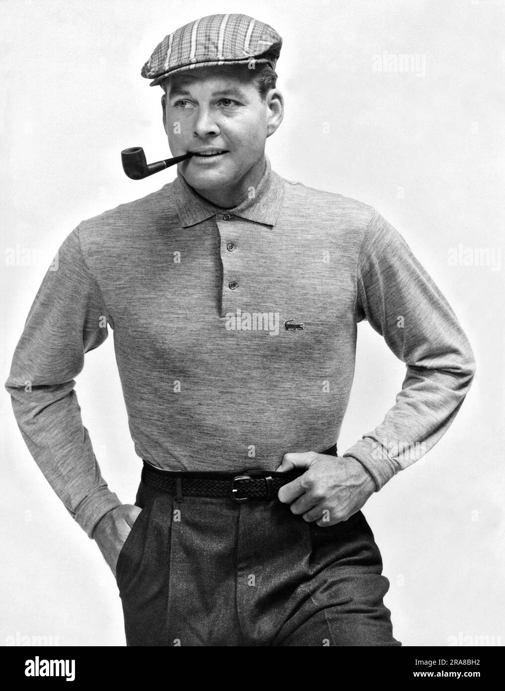 États-Unis: c. 1954 Un homme à la mode fume un tuyau et porte une tenue de sport décontractée d'un chandail Lacoste et d'une casquette de golf plate. Banque D'Images