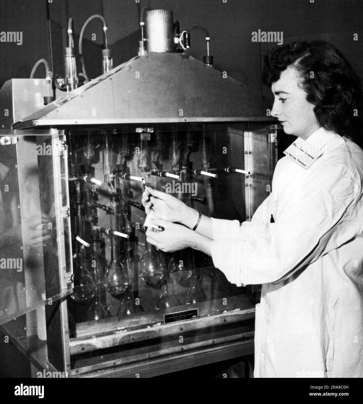 New York, New York. 9 janvier 1958 Joan Johnson, de la société d'essais des États-Unis, donne à la machine à fumer une lumière pour chacune de ses dix cigarettes. La fumée est recueillie dans des flacons pour l'analyse chimique de la teneur en nicotine et en goudron pour le nouveau filtre à cigarettes de la société Phiip Morris. Banque D'Images