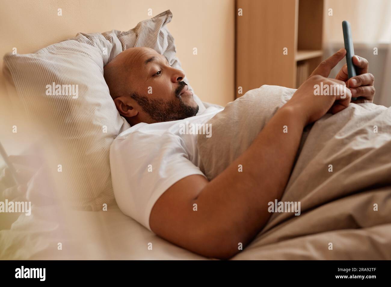 Vue latérale d'un homme adulte noir utilisant un smartphone dans le lit à la maison le matin d'un repos Banque D'Images