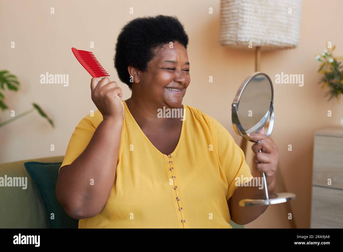 Gaie femme afro-américaine mature avec une brosse à cheveux en plastique rouge regardant dans le miroir tout en prenant soin de ses cheveux le matin Banque D'Images