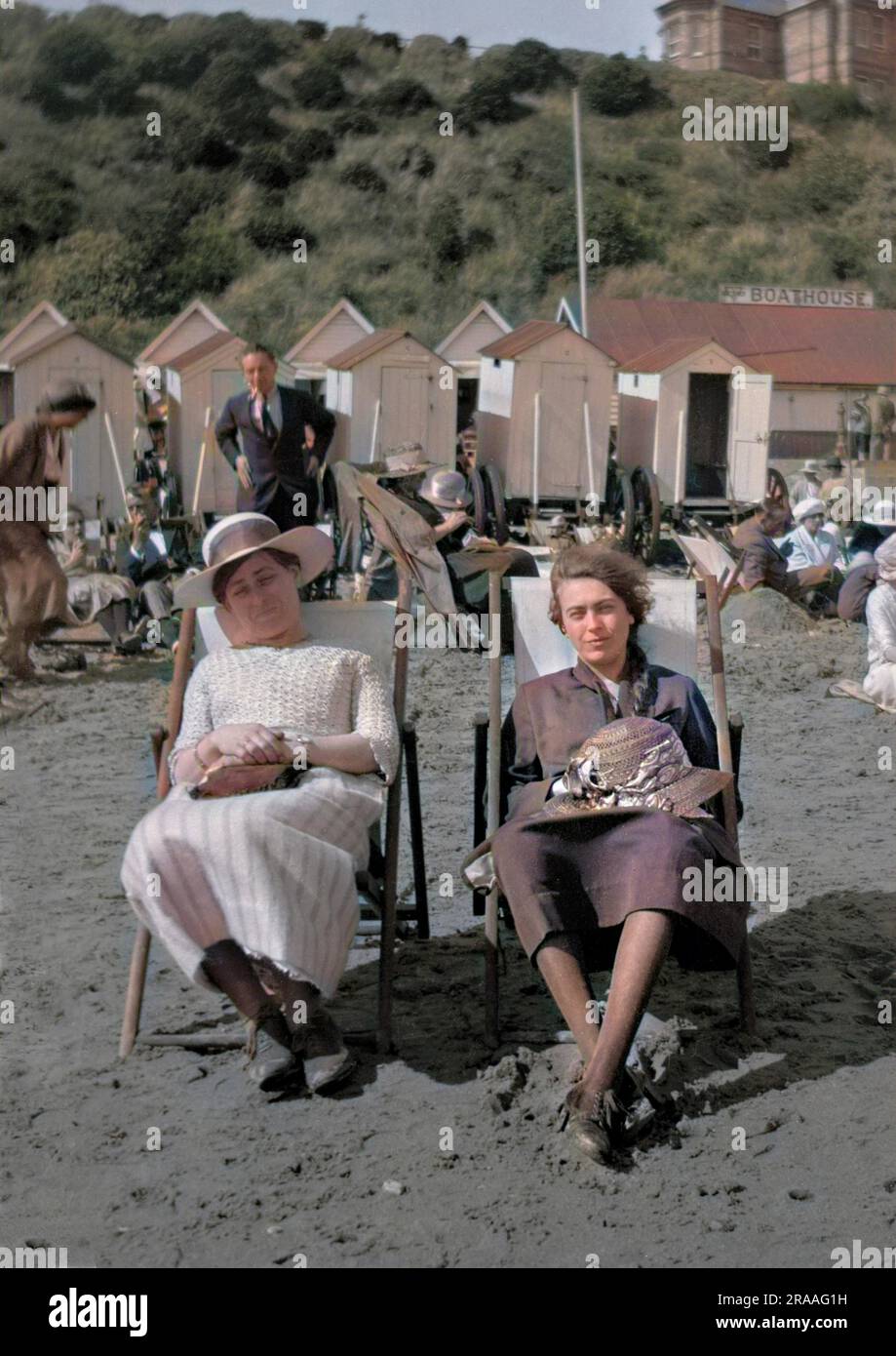 Deux jeunes femmes assises sur une plage dans des transats, avec des cabines de bain en arrière-plan. Banque D'Images