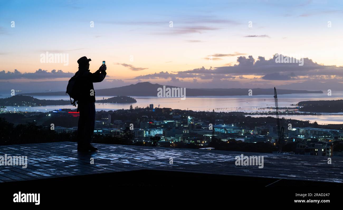 Homme qui prend des photos à l'aube à l'aide d'un smartphone au sommet du Mont Eden. Rangitoto Island au loin. Auckland. Banque D'Images