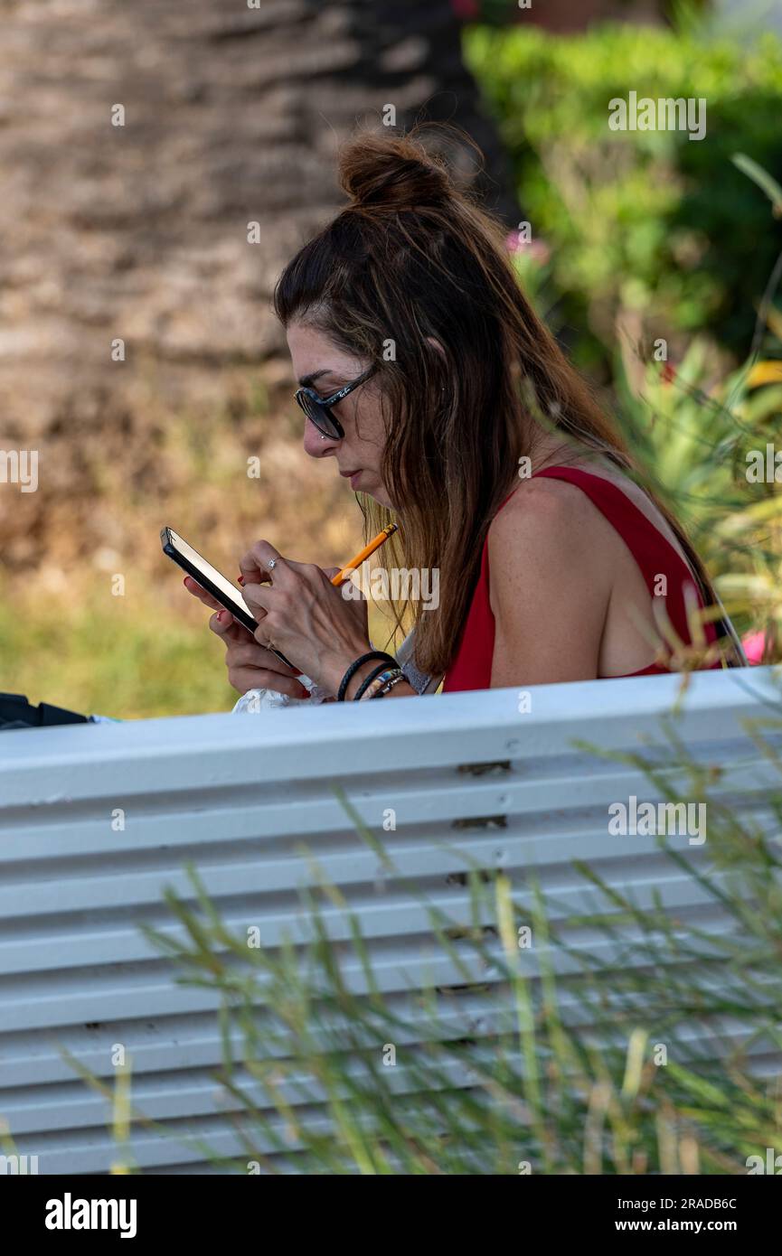 jeune femme absorbée en regardant un écran de smartphone portant des lunettes de soleil. jeune femme utilisant smartphone ou téléphone portable assis sur un banc de parc Banque D'Images