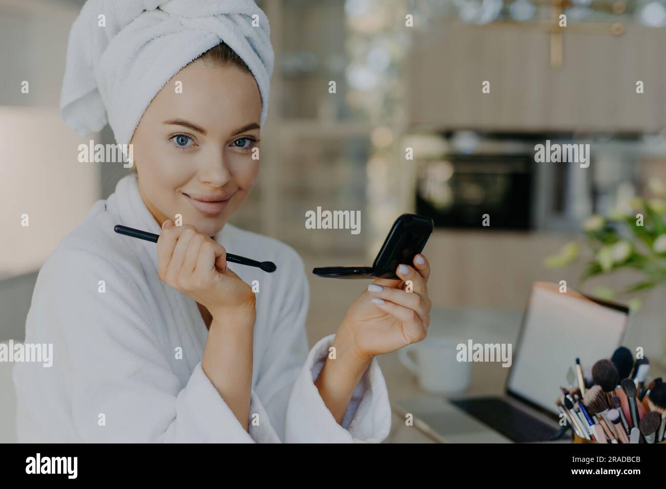 Une jeune femme applique du maquillage, pose dans un peignoir, avec un pinceau, dans un appartement moderne. Banque D'Images