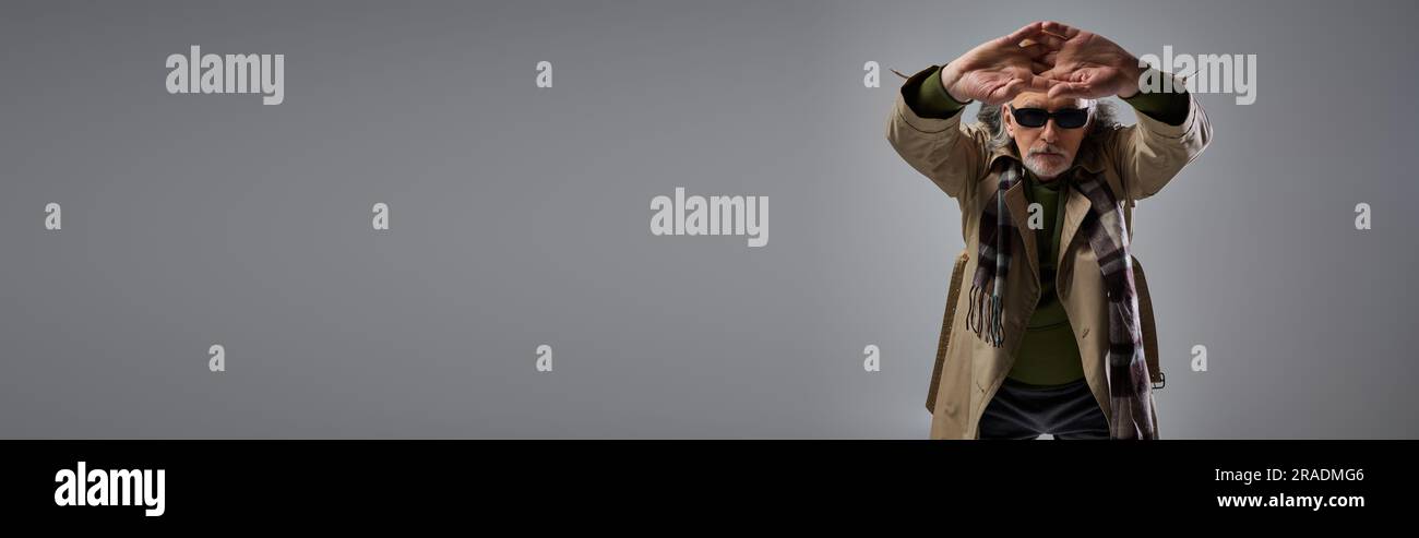 style hipster tendance homme en lunettes de soleil sombres et trench-coat beige posant avec les mains tendues sur fond gris, âge élégant, mode s. Banque D'Images