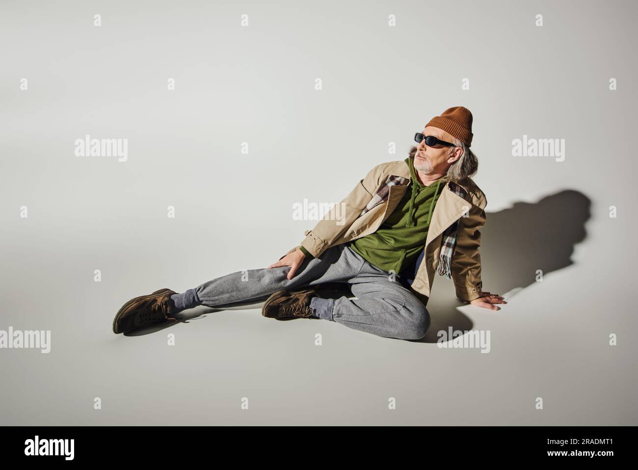 sur toute la longueur, modèle masculin senior tendance, assis et regardant loin sur fond gris avec ombre, lunettes de soleil sombres, bonnet beanie, trench-coat beige, Banque D'Images