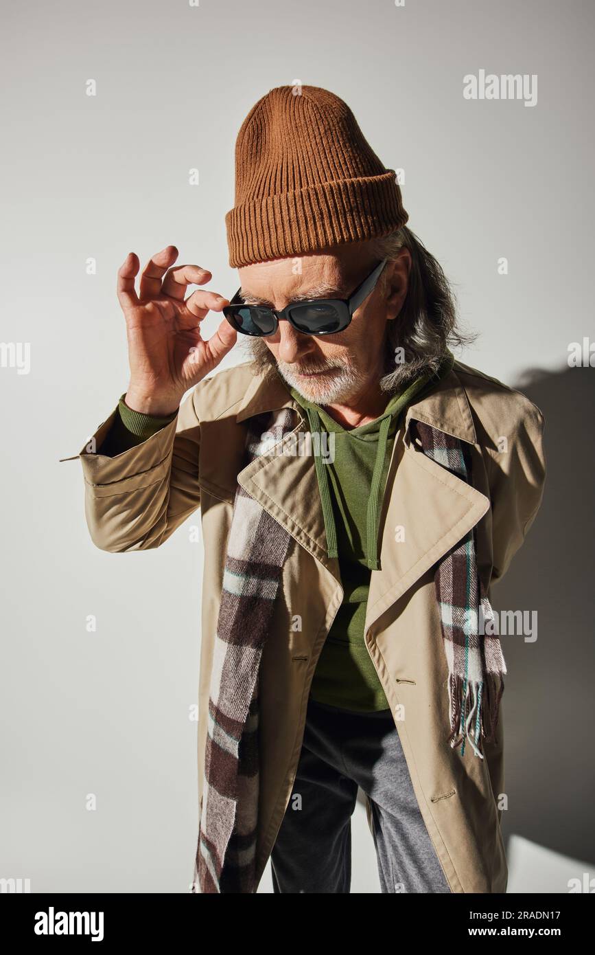 homme barbu et senior en bonnet beanie echarpe a motif ecossais et trench beige ajustement des lunettes de soleil sombres sur fond gris avec ombre vetements taille basse givre Photo Stock