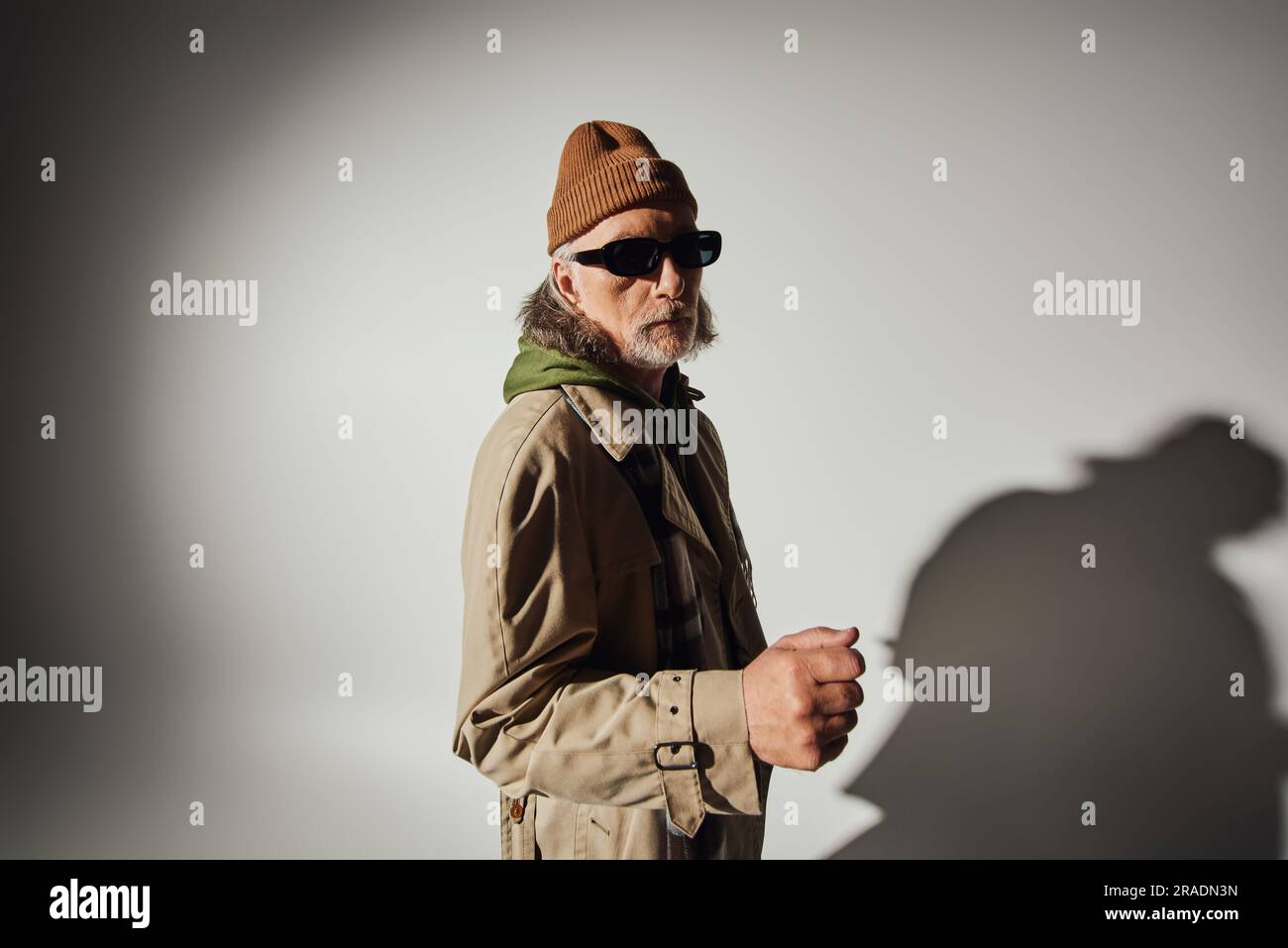 modèle senior masculin en lunettes de soleil sombres et vêtements décontractés élégants regardant l'appareil photo sur fond gris avec ombre, bonnet beanie, trench-coat beige, tissu écossais Banque D'Images
