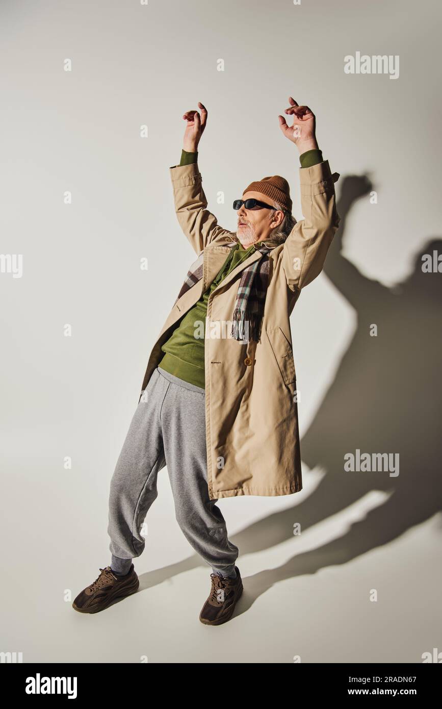 pleine longueur de style hippster expressif homme senior en lunettes de soleil sombres, bonnet beanie, trench-coat beige et écharpe à carreaux posant avec les mains levées sur le gris Banque D'Images