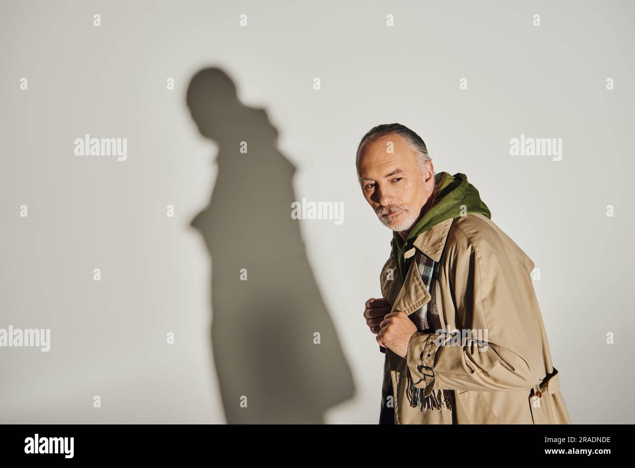 modèle masculin senior et réfléchi à capuche vert, écharpe à motif écossais et trench beige debout sur fond gris avec ombre et vue à l'extérieur, personnel Banque D'Images